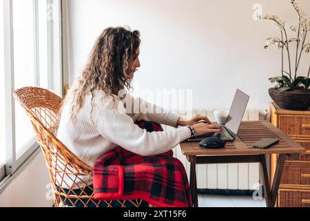 Seitenansicht einer jungen Freiberuflerin, die mit Laptop am Tisch sitzt und Informationen schreibt, während sie an einem neuen Projekt im modernen Zuhause arbeitet Stockfoto