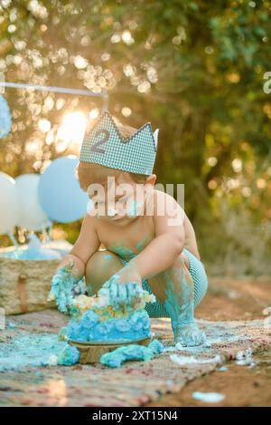 Ein süßer Kleinkind sitzt draußen und feiert seinen zweiten Geburtstag mit einem unordentlichen Kuchenschlag, gekleidet in einer Windel und einer Papierkrone, in die er fröhlich hineingräbt Stockfoto