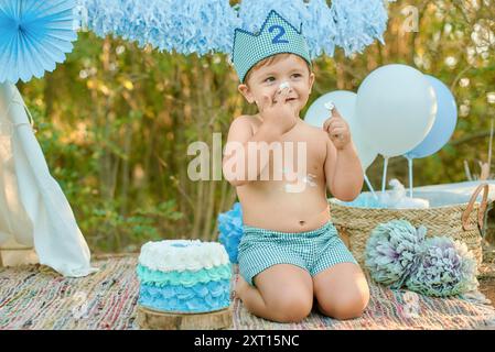 Wunderbares Bild eines Kleinkindes mit Partyhut, der seinen zweiten Geburtstagskuchen zwischen Dekorationen, Ballons und Waldkulisse genießt Stockfoto