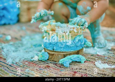 Ein junges Kleinkind zertrümmert bei einer Feier im Freien einen blauen Milchkuchen und verschmiert sich mit Glasur über sich und die Umgebung Stockfoto