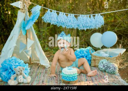 Ein kleiner Junge genießt seinen zweiten Geburtstag mit einem festlichen Kuchen im Freien, gekleidet mit einer karierten Krone und passendem Windelbezug, er genießt das Essen Stockfoto