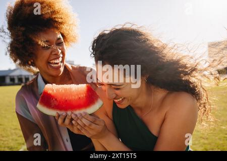 Zwei beste Freundinnen lachen und genießen einen sonnigen Sommertag im Park mit einer saftigen Wassermelonenscheibe. Stockfoto