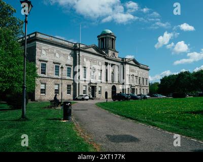 Ein großes historisches Gebäude steht stolz in einem lebhaften grünen Park, der sich unter dem klaren blauen Himmel sonnt. Besucher schlendern entlang des Weges und genießen die Stockfoto