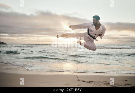 Karate, Mann und Jump Kick am Strand für Bewegung, Körpergesundheit und Taekwondo im Freien. Kämpfer, Fliegen und Kampfkunst auf dem Ozean für Fitness Stockfoto