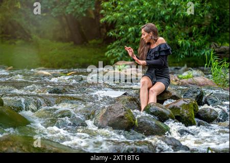 Eine Frau benutzt einen Computer, während sie auf einem Felsen im Fluss sitzt, Laptop fällt ins Wasser, Unfall Stockfoto