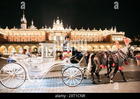 Krakau, Polen. Pferde in einer altmodischen Kutsche, die in der Nähe des Tuchhalle-Gebäudes am Altstädter Ring in der Sommernacht unterwegs sind. Stockfoto