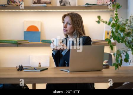 Durchdachte Geschäftsfrau trinkt Kaffee während der Arbeit im Büro mit Laptop, Multitasking und Arbeit Stockfoto