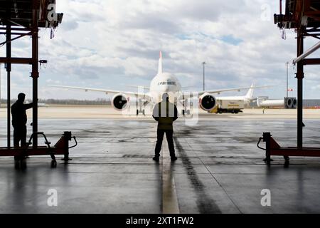 Das Mitglied der Bodenbesatzung steht in einem Flugzeughangar gegenüber einem geparkten Verkehrsflugzeug, während ein anderes Flugzeug im Hintergrund sichtbar ist. Bola02491 Copyrig Stockfoto