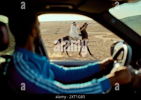 Blick aus dem Innenraum eines Autos auf eine Person, die auf einem Kamel durch eine Wüstenlandschaft reitet, durch die Windschutzscheibe mit dem Fahrer in Teilsicht gesehen. Bola0253 Stockfoto