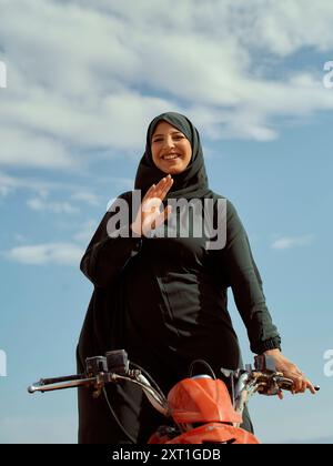 Lächelnde Frau in einem Hijab, die einen roten Roller vor einem blauen Himmel fährt. Bola02569 Copyright: XConnectxImagesx Stockfoto