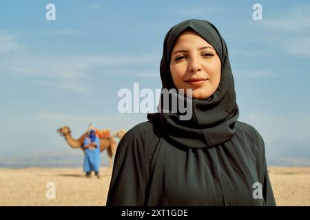 Eine lächelnde Frau in einem Hijab steht in einer Wüstenlandschaft mit einem Kamel und einer Person in blauer traditioneller Kleidung im Hintergrund. Bola02557 Copyrig Stockfoto