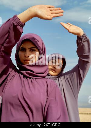 Zwei Frauen in lila Hijabs posieren vor einem klaren Himmel mit symmetrisch erhobenen Armen. Bola02560 Copyright: XConnectxImagesx Stockfoto