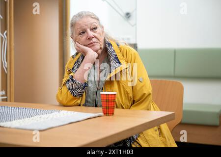 Eine ältere Frau in einer gelben Jacke sitzt mit nachdenklichem Blick an einem Tisch und legt ihren Kopf auf die Hand. Maulwurf Stockfoto