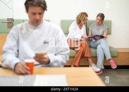 Ein junger Mann überprüft sein Handy im Vordergrund, während zwei Frauen zusammen sitzen und in einer Wartezeit eine Zeitschrift lesen Stockfoto