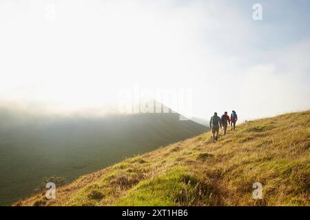 Eine Gruppe von Wanderern geht im Morgennebel einen grünen, grasbewachsenen Hügel hinauf, mit einem klaren Himmel über und einem Berghang im Hintergrund. Phhi00062 Copyright Stockfoto