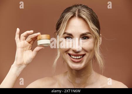 Eine junge Frau mit Vitiligo lächelt hell, während sie ein Glas Creme vor einem beigen Hintergrund hält. Stockfoto