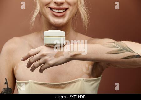 Eine junge Frau mit Vitiligo lächelt und hält ein Cremeglas in der Hand vor beigem Hintergrund. Stockfoto