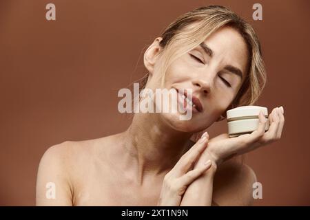 Eine junge Frau mit Vitiligo hält einen Cremebecher, ihre Augen sind zufrieden geschlossen, vor einem beigen Hintergrund. Stockfoto