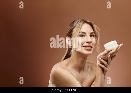 Eine junge Frau mit Vitiligo lächelt zuversichtlich, während sie ein Glas Creme vor einem beigefarbenen Hintergrund hält. Stockfoto