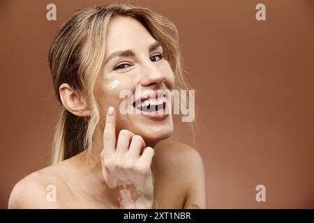 Eine junge Frau mit Vitiligo lächelt, als sie Creme auf ihr Gesicht aufträgt, vor einem beigen Hintergrund. Stockfoto