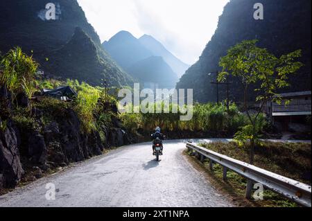 Ein Motorradfahrer fährt auf einer gewundenen Straße, umgeben von üppigem Grün und hohen Bergen in einem ruhigen Tal. HaGiang Vietnam rist00109 Copyright: XCO Stockfoto