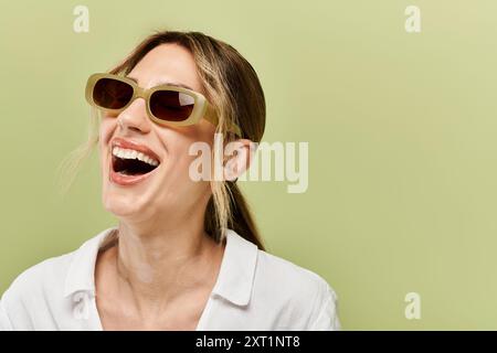 Eine junge Frau mit Vitiligo lächelt hell, während sie eine Sonnenbrille und ein weißes Sommerkleid vor grünem Hintergrund trägt. Stockfoto