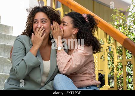 Zwei Frauen teilen sich einen Moment des Lachens und Flüsters auf den Stufen im Freien. Panc05404 Copyright: XConnectxImagesx Stockfoto