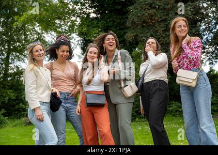 Eine Gruppe von sechs lächelnden Frauen, die draußen auf einem grasbewachsenen Gebiet stehen, jede in lässiger bis eleganter Freizeitkleidung gekleidet und einen gemeinsamen Moment unter einem Clo genießen Stockfoto