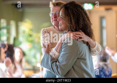 Ältere Frau mit Brille, die eine jüngere Frau mit lockigen Haaren umschließt, drinnen mit verschwommenen Menschen im Hintergrund. Panc05587 Copyright: XConnectxImag Stockfoto
