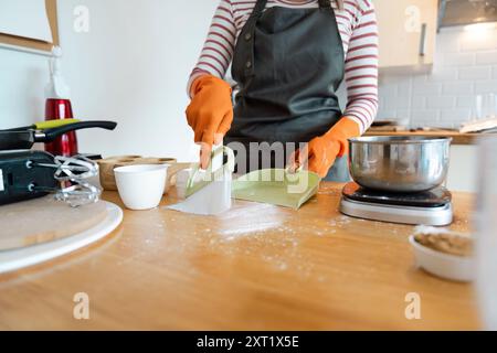 Frau, die Küchenzeile mit Staubpfanne und Pinsel reinigt, trägt orangefarbene Handschuhe in einem modernen Zuhause Stockfoto