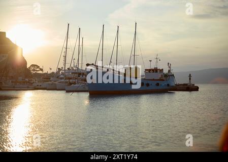Blick am frühen Morgen auf einen ruhigen Hafen mit einem blauen Boot, das neben einem Pier liegt, mit sanftem Sonnenlicht, das vom ruhigen Wasser glitzert. Hydra Griechenland Bola0 Stockfoto