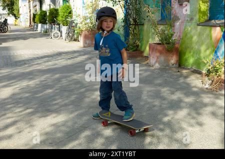 Ein kleiner Junge steht selbstbewusst auf einem Skateboard, trägt einen Helm und legere Kleidung mit einer sonnendurchfluteten Graffiti-Wand im Hintergrund. Bola02710 Copyright Stockfoto