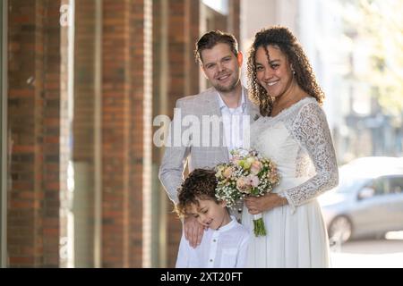 Ein Paar in Hochzeitskleidung und ein Kind lächelt auf einer sonnigen Straße, die Frau hält einen Blumenstrauß. Panc05769 Copyright: XConnectxIm Stockfoto