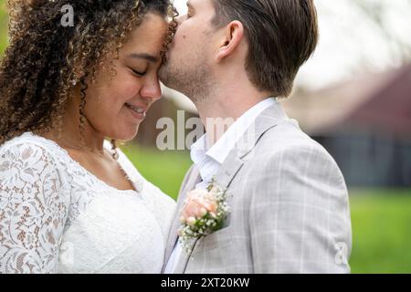 Liebevolles Paar in Hochzeitskleidung, das einen zärtlichen Moment mit angreifenden Stirnen und geschlossenen Augen teilt und draußen mit Grün im Hintergrund steht. PA Stockfoto