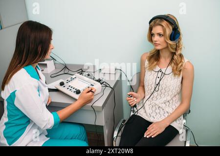 Eine junge Frau nimmt an einem Hörtest Teil, während sie Kopfhörer trägt und von einem medizinischen Fachpersonal in einer klinischen Umgebung geleitet wird. Stockfoto