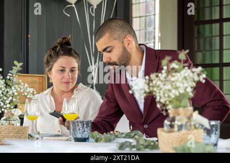 Ein professioneller Mann im Anzug und eine Frau in lässiger Kleidung, die gemeinsam ein mobiles Gerät an einem elegant gedeckten Tisch mit Blumen und Getränken untersuchen. Panc058 Stockfoto