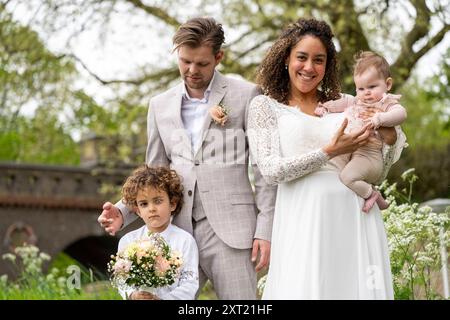 Eine in formeller Kleidung gekleidete Familie posiert zusammen in einem Park, der Vater steht hinter einem Kleinkind mit Blumen und die Mutter hält einen Infan Stockfoto