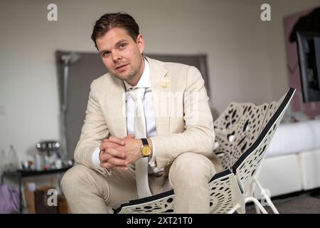 Gut gekleideter Mann in einem beigefarbenen Anzug, der drinnen auf einem modernen Stuhl sitzt und selbstbewusst mit einem leichten Lächeln auf die Kamera blickt. Panc05941 Copyright: XConne Stockfoto
