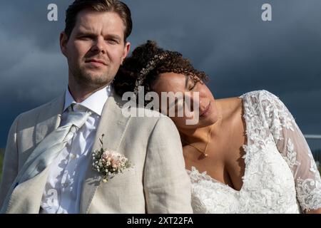 Die Braut lehnt sich auf die Schulter des Bräutigams unter einem dramatischen bewölkten Himmel, beide in eleganter Hochzeitskleidung. Panc05985 Copyright: XConnectxImagesx Stockfoto