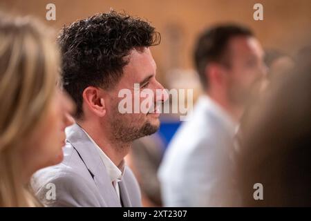 Seitenprofil eines lächelnden Mannes mit lockigem Haar, der an einer Veranstaltung teilnimmt, wobei andere Teilnehmer im Vorder- und Hintergrund verschwimmen. Panc06031 Copyright: Stockfoto