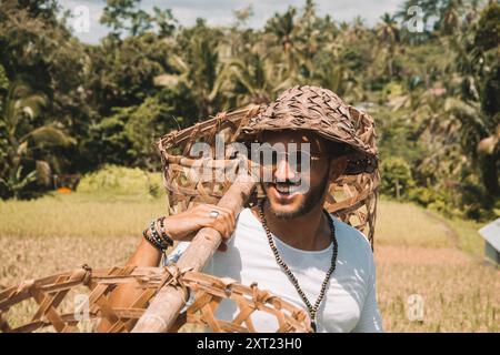 Lächelnder junger Mann auf einem Bali-Reisfeld, der einen Strohhut trägt und ein traditionelles Werkzeug hält, um den Reis zu sammeln Stockfoto