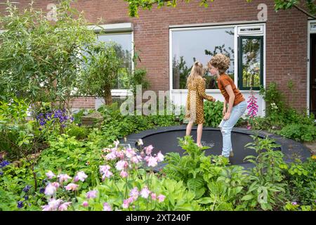 Zwei Kinder spielen auf einem Gartentrampolin in der Nähe eines Backsteinhauses mit üppigem Grün. Panc06115 Copyright: XConnectxImagesx Stockfoto