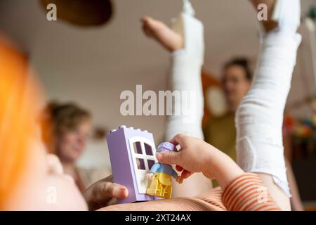 Ein Kind spielt mit einem Spielzeughaus aus Plastik in einem gemütlichen Zuhause, während sich Erwachsene im Hintergrund mischen. Panc06155 Copyright: XConnectxImagesx Stockfoto