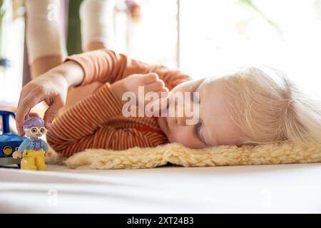 Ein Kind mit Beinabschlüssen spielt mit Spielzeug auf einer weißen Decke drinnen, mit Plüschspielzeug im Hintergrund und Blick durch das Fenster. Pfannen06143 C Stockfoto