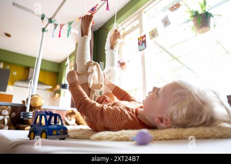 Ein Kind mit Beinabschlüssen spielt mit Spielzeug auf einer weißen Decke drinnen, mit Plüschspielzeug im Hintergrund und Blick durch das Fenster. Pfannen06145 C Stockfoto