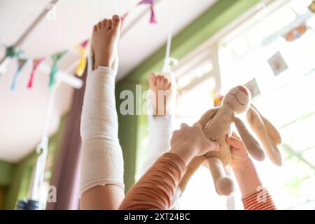 Ein Kind mit Beinabschlüssen spielt mit Spielzeug auf einer weißen Decke drinnen, mit Plüschspielzeug im Hintergrund und Blick durch das Fenster. Pfannen06148 C Stockfoto