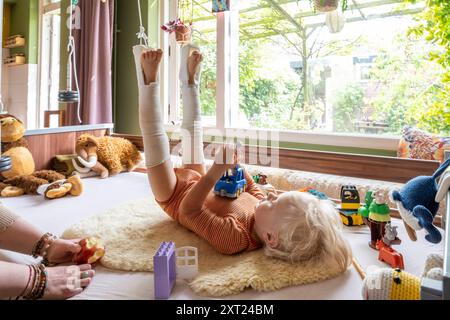 Ein Kind mit Beinabschlüssen spielt mit Spielzeug auf einer weißen Decke drinnen, mit Plüschspielzeug im Hintergrund und Blick durch das Fenster. Pfannen06141 C Stockfoto