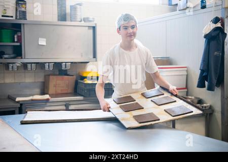 Banketbakkerij Ein lächelnder Bäcker in einem weißen T-Shirt trägt ein großes Tablett mit frischen Brownies in einer professionellen Küche. Molenhoek Nederland Panc06194 Kopie Stockfoto