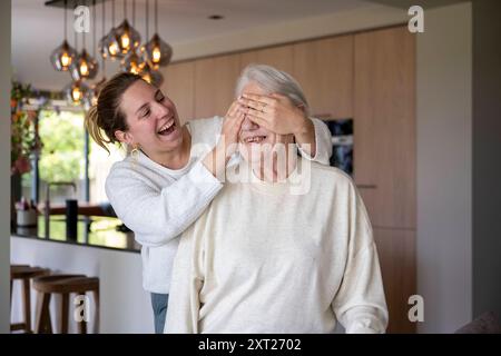 Eine fröhliche junge Frau bedeckt die Augen einer älteren Frau von hinten, die beide in einer warm beleuchteten modernen Küche lachen. Panc06377 Copyright: XConnectxIm Stockfoto