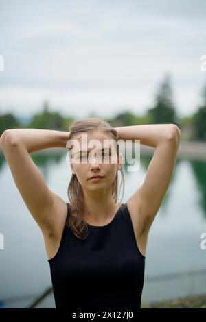 Die junge Frau in einem schwarzen Dach steht selbstbewusst mit den Händen hinter dem Kopf vor einem verschwommenen See-Hintergrund. Bola02772 Copyright: XConnectxImagesx Stockfoto